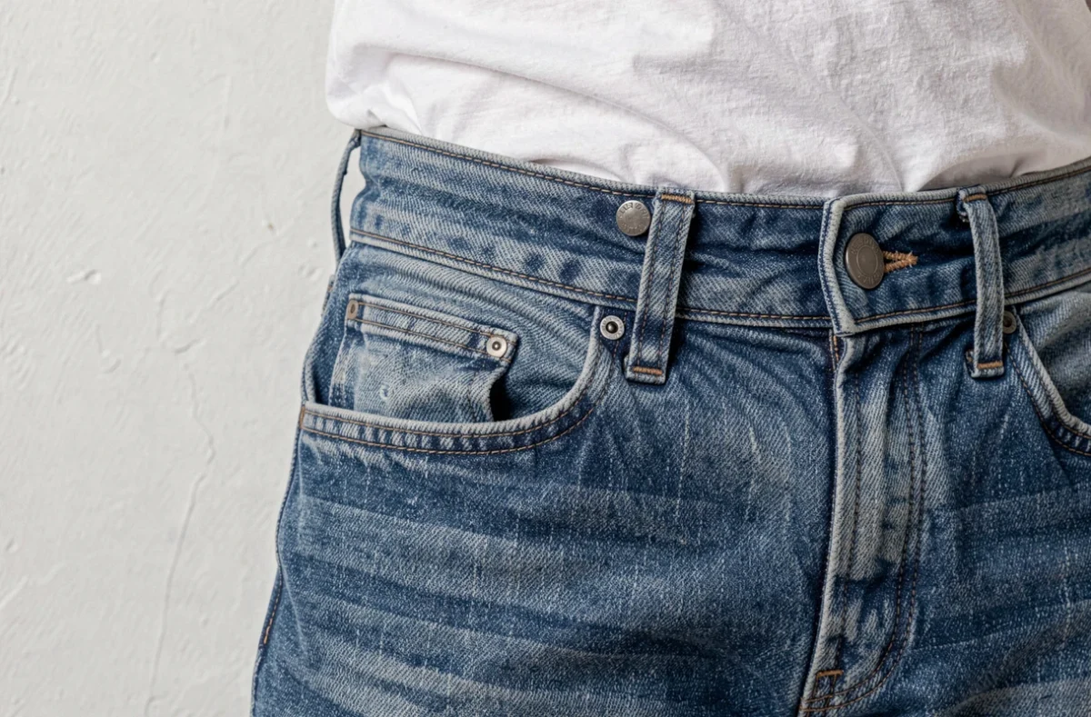 Bouton de jean passé dans le passant de ceinture adjacent pour resserrer le tour de taille sans accessoire supplémentaire